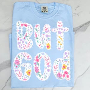 Op de afbeelding: Lichtblauw T-shirt met de woorden "BUT GOD" in witte, met bloemen gevulde letters. De letters zijn omlijnd met een witte, geschulpte rand. Het shirt is gemaakt van een zacht materiaal en heeft een ronde hals.