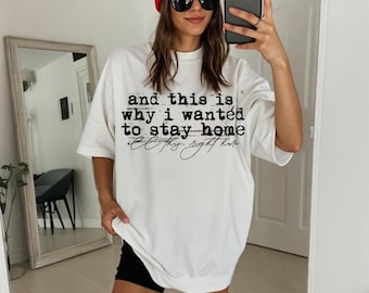 May include: White oversized t-shirt with the text "and this is why I wanted to stay home" in black. The shirt is paired with black shorts and a red beanie. The person is wearing sunglasses and holding a phone.
