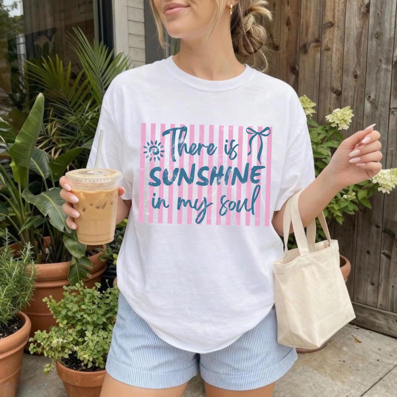 Puede incluir: Camiseta blanca con el texto "There is sunshine in my soul" en azul y rosa. El texto est&aacute; sobre un fondo de rayas rosas. La persona lleva pantalones cortos a rayas azules y blancas y sostiene una bebida.