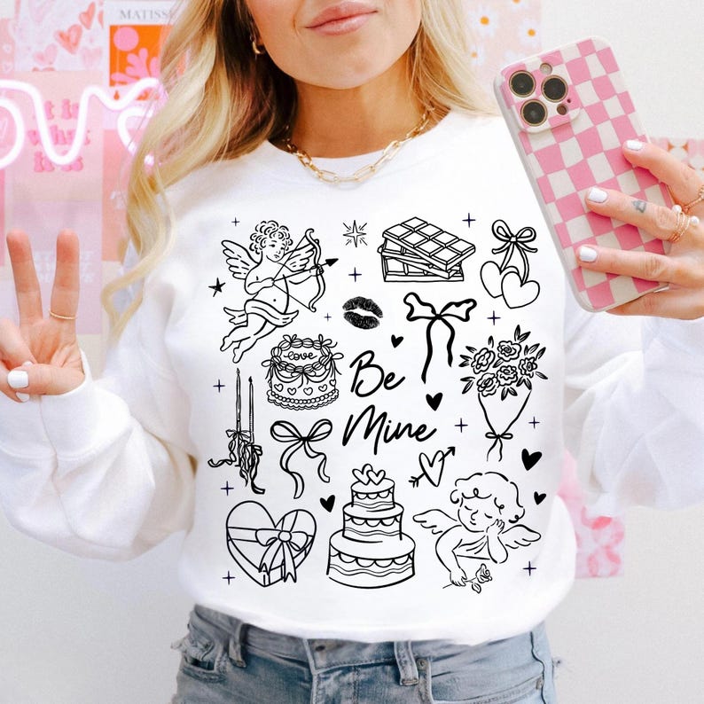 May include: White sweatshirt with black line art illustrations of Valentine's Day themed images, including a cupid, cake, chocolate, and the words "Be Mine". The person is holding a pink and white checkered phone case.