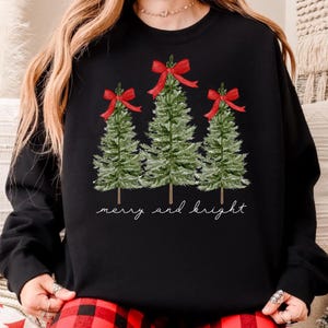 May include: Black sweatshirt featuring three green Christmas trees with red bows. The words "merry and bright" are written in a cursive font below the trees. A festive holiday design.