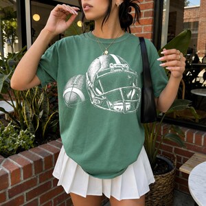 May include: A sage green short-sleeve t-shirt featuring a white graphic print of an American football helmet and a football. The shirt is paired with a white pleated skirt and a black shoulder bag, with a gold chain necklace worn over the shirt.