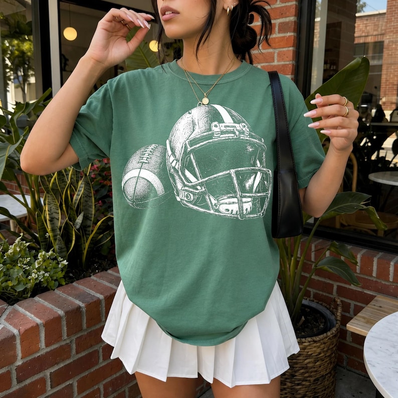 May include: A sage green short-sleeve t-shirt featuring a white graphic print of an American football helmet and a football. The shirt is paired with a white pleated skirt and a black shoulder bag, with a gold chain necklace worn over the shirt.