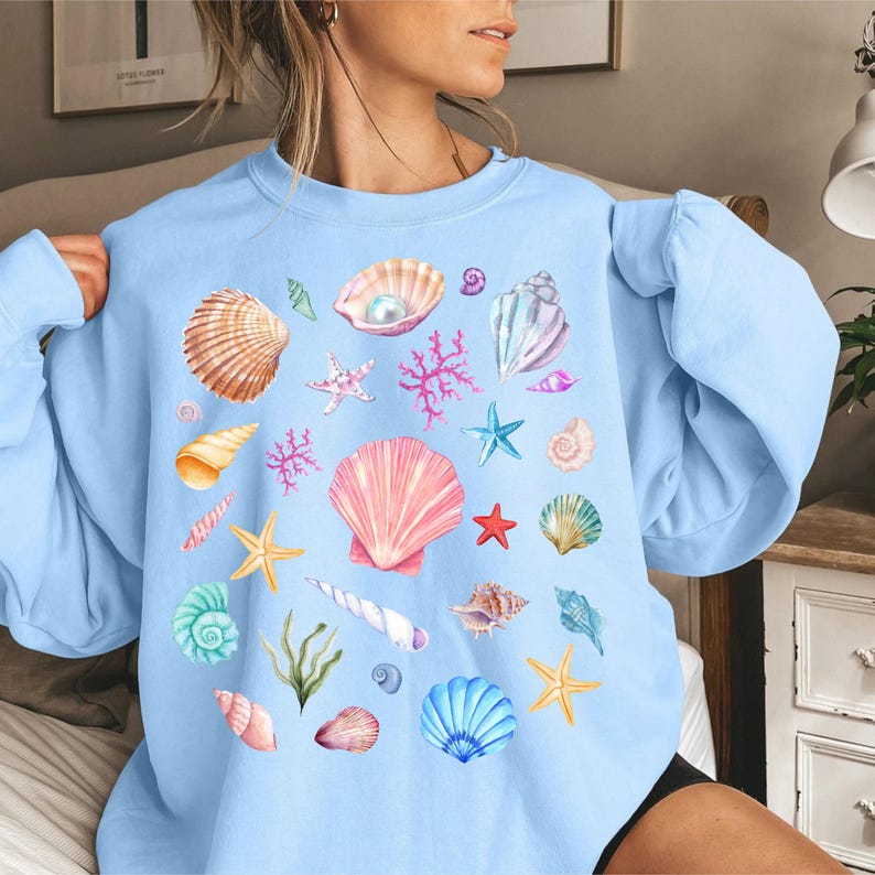 May include: A light blue sweatshirt with a colorful seashell print. The print features various seashells, starfish, and coral in shades of pink, blue, green, and white.