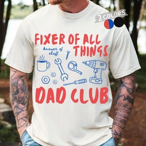Op de afbeelding: Crèmekleurig T-shirt met de rode tekst "FIXER OF ALL THINGS" en "DAD CLUB". Het shirt heeft blauwe illustraties van gereedschap en een koffiekopje. De zin "knower of stuff" is ook opgenomen.