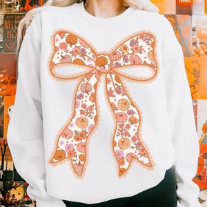 May include: White sweatshirt featuring a large bow design. The bow is decorated with pumpkins and flowers in shades of orange, pink, and peach. The bow is outlined with a peach-colored trim and lace detailing. The sweatshirt is a fall-themed item.