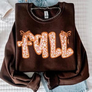 May include: A dark brown sweatshirt with the word "FALL" in a retro font. The letters are filled with a floral pattern of orange and pink flowers and outlined in orange. Two orange bows are on the letters.