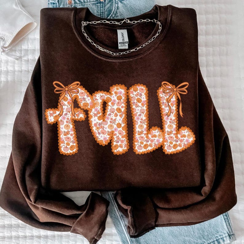 May include: A dark brown sweatshirt with the word "FALL" in a retro font. The letters are filled with a floral pattern of orange and pink flowers and outlined in orange. Two orange bows are on the letters.