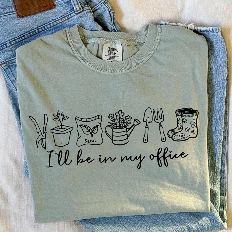 Puede incluir: Camiseta verde salvia con un gr&aacute;fico negro de herramientas de jardiner&iacute;a y el texto "I'll be in my office." El dise&ntilde;o incluye unas tijeras de podar, una planta en maceta, un sobre de semillas, una regadera, una paleta, un tenedor y una bota de agua.