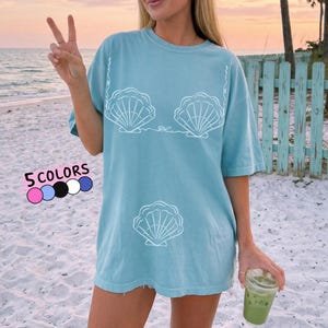 May include: A light blue t-shirt with a white seashell design. The shirt features a crew neck and short sleeves. The text "5 COLORS" is displayed with color swatches. The shirt is worn on a beach.