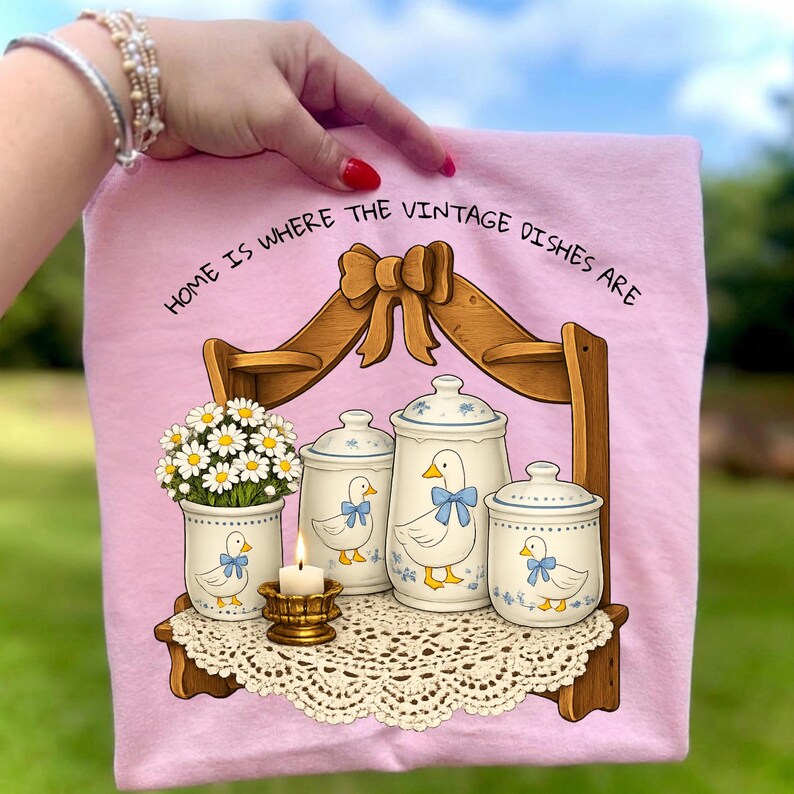 Op de afbeelding: Roze decoratieve handdoek met de tekst "HOME IS WHERE THE VINTAGE DISHES ARE." De handdoek heeft een plank met potten, een vaas met madeliefjes en een kaars. De potten en vaas hebben een eendenontwerp.
