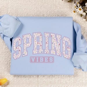 May include: A light blue sweatshirt with the word "SPRING" in large, floral-patterned letters, and the word "VIBES" below. The floral pattern includes pink, purple, and white flowers. The sweatshirt is folded.