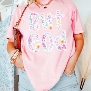 Op de afbeelding: Roze T-shirt met de tekst "BUT GOD" in een bloemenontwerp. De letters zijn gevuld met verschillende bloemen in roze, paarse, gele en groene tinten. Het shirt heeft korte mouwen.