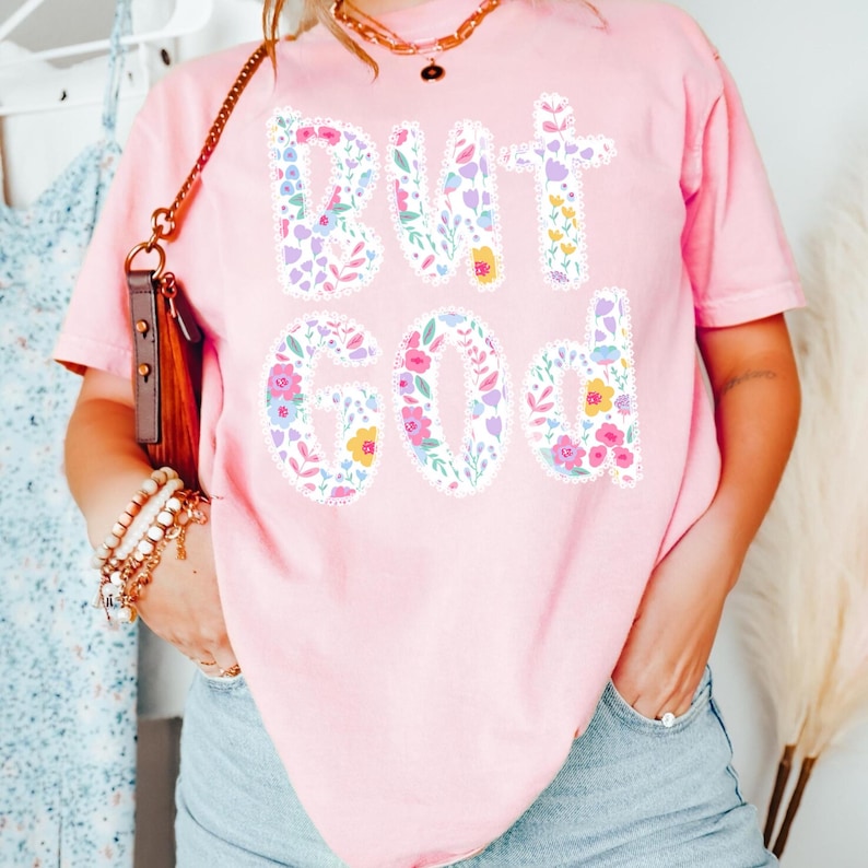 Op de afbeelding: Roze T-shirt met de tekst "BUT GOD" in een bloemenontwerp. De letters zijn gevuld met verschillende bloemen in roze, paarse, gele en groene tinten. Het shirt heeft korte mouwen.