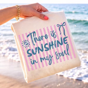 Puede incluir: Un bolso tote crema con rayas rosas y blancas y el texto turquesa "There is SUNSHINE in my soul". El bolso es sostenido por una persona con una pulsera dorada y esmalte de u&ntilde;as rosa, con el mar de fondo.