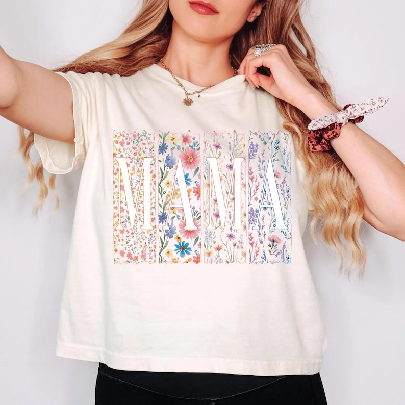 May include: Cream-colored t-shirt with the word "MAMA" in white block letters. Each letter is filled with a different floral pattern in shades of pink, blue, yellow, and purple. The shirt has short sleeves.