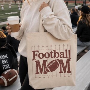 May include: Beige tote bag with the words "Football Mom" in a distressed brown font, along with a football graphic. The bag has a football field design on the top and bottom. A football and a coffee cup are also visible.
