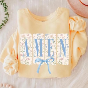 May include: A light yellow sweatshirt with the word "AMEN" in light blue block letters. The letters are set against a floral background with a light blue bow at the bottom. The sweatshirt is folded.