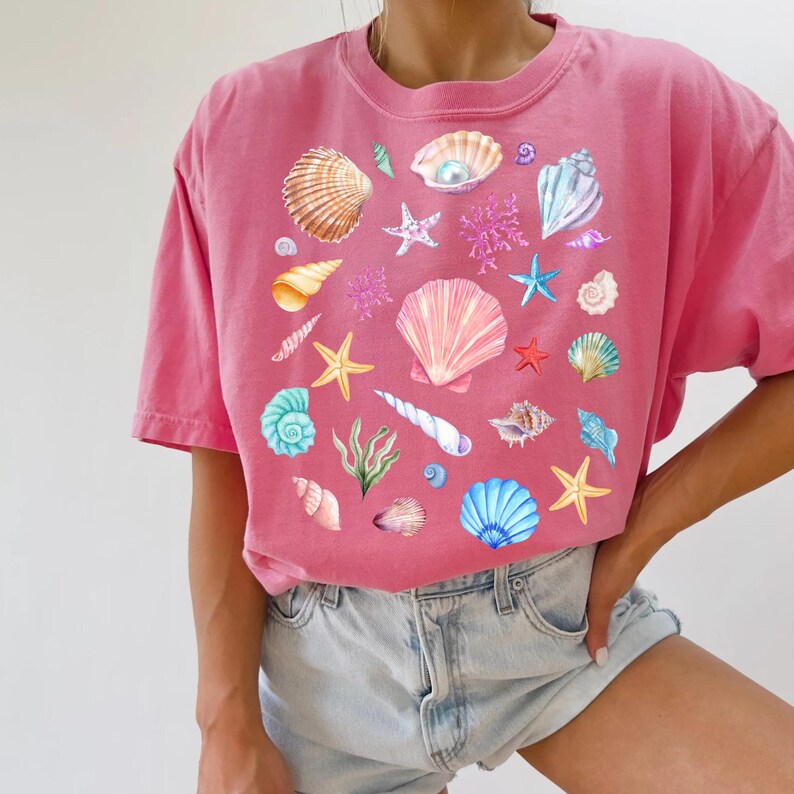 May include: A pink t-shirt with a colorful seashell design. The design features various seashells, starfish, and coral in shades of pink, blue, green, and white.