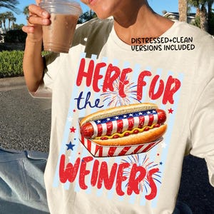 Op de afbeelding: Crèmekleurig T-shirt met een afbeelding van een hotdog in een patriottisch ontwerp. De tekst "HERE FOR the WEINERS" is rood. Het shirt bevat de tekst "DISTRESSED+CLEAN VERSIONS INCLUDED".