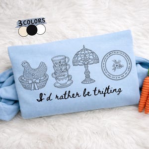 May include: Light blue graphic tee with black line art illustrations of a hen in a basket, stacked teacups, a lamp, and a floral plate. The text "I'd rather be thrifting" is below the illustrations. The top left corner has a color palette.