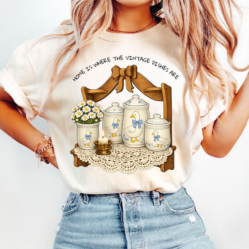 Op de afbeelding: Cr&egrave;mekleurig T-shirt met een grillige illustratie van een houten plank met vintage potten, een vaas met bloemen en een kaars. De potten hebben eendenontwerpen en blauwe accenten. De tekst "HOME IS WHERE THE VINTAGE DISHES ARE" staat er boven.