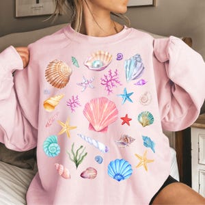 May include: A pink sweatshirt with a colorful seashell print. The print features various seashells, starfish, and coral in shades of pink, blue, green, and white.