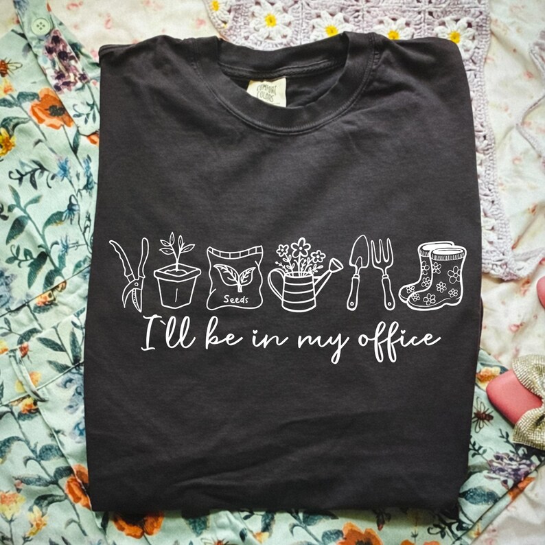 Puede incluir: Camiseta negra con dibujos lineales blancos de herramientas de jardiner&iacute;a y plantas. El texto "I'll be in my office" est&aacute; debajo. La camiseta est&aacute; doblada sobre una superficie con estampado floral.