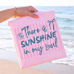 Puede incluir: Una toalla de playa rosa con rayas verticales y el texto "There is SUNSHINE in my soul" en color turquesa. La toalla se sostiene sobre un fondo de playa, con el oc&eacute;ano visible al fondo.
