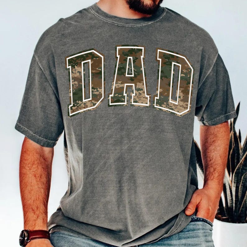 May include: A gray t-shirt with a camouflage print of the word "DAD" in white outlined letters.