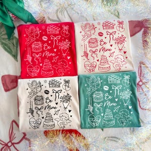 May include: Four t-shirts in red, cream, and green, each with a Valentine's Day design. The design includes cherubs, cakes, bows, and the words "Be Mine". The shirts are arranged in a square.