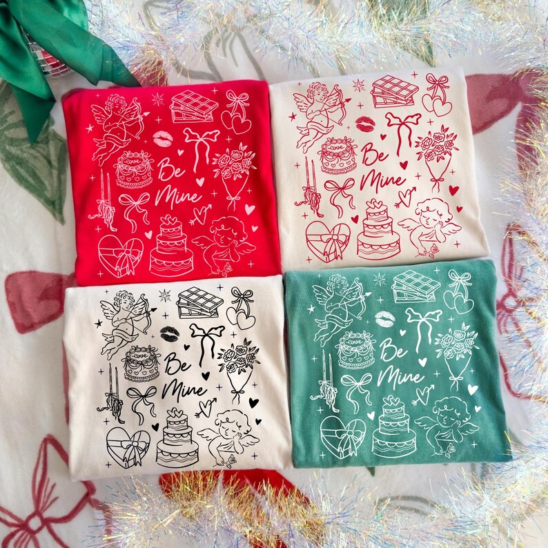 May include: Four t-shirts in red, cream, and green, each with a Valentine's Day design. The design includes cherubs, cakes, bows, and the words "Be Mine". The shirts are arranged in a square.