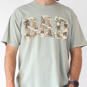 May include: A light green t-shirt with a camouflage print "DAD" graphic.