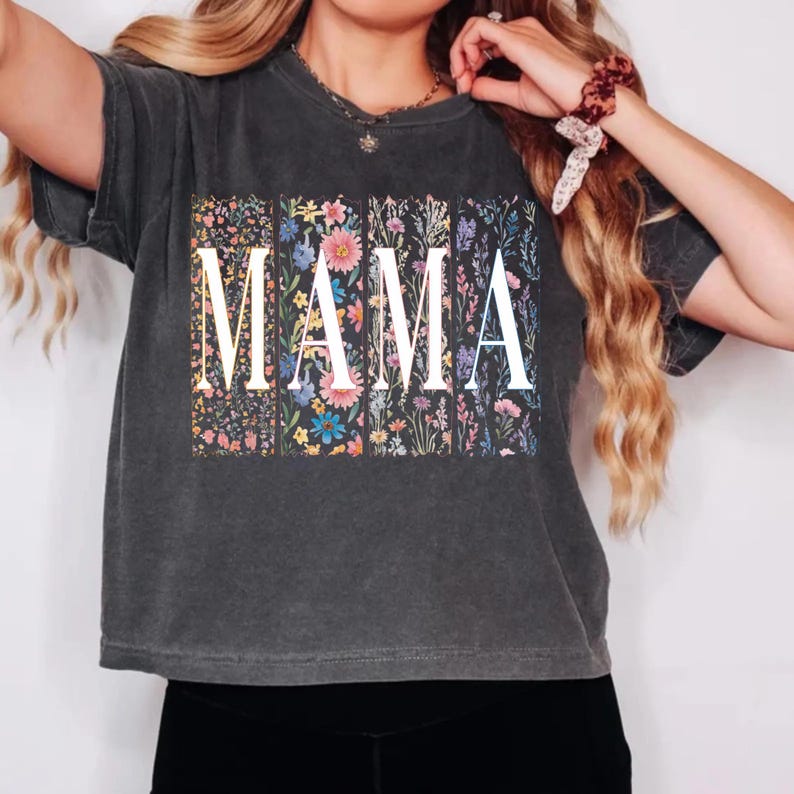May include: Dark gray t-shirt with the word "MAMA" in large white letters. The letters are superimposed over a floral design with colorful flowers. The shirt has short sleeves and a crew neck.