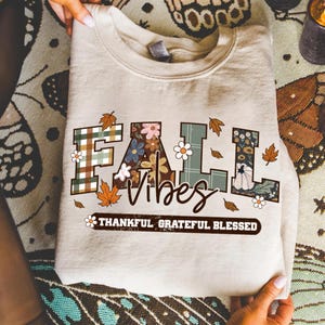 May include: A beige sweatshirt with the words "FALL VIBES" in a decorative font. The letters are filled with floral and plaid patterns. The words "THANKFUL GRATEFUL BLESSED" are printed below. Autumn leaves and flowers are scattered around the text.