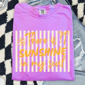 Puede incluir: Una camiseta lila Comfort Colors con el texto "There is Sunshine in my Soul" en amarillo. El texto est&aacute; sobre un fondo de rayas verticales crema. La camiseta est&aacute; doblada y colocada sobre una superficie estampada.