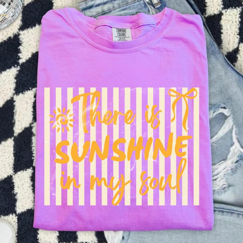 Puede incluir: Una camiseta lila Comfort Colors con el texto "There is Sunshine in my Soul" en amarillo. El texto est&aacute; sobre un fondo de rayas verticales crema. La camiseta est&aacute; doblada y colocada sobre una superficie estampada.