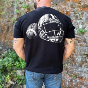 May include: A black short-sleeved t-shirt featuring a white graphic print on the back. The design displays a detailed illustration of a football helmet positioned next to a football, both rendered in a sketch-like style with high contrast.
