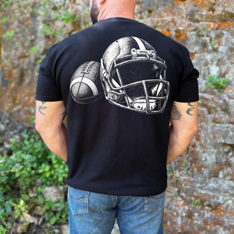 May include: A black short-sleeved t-shirt featuring a white graphic print on the back. The design displays a detailed illustration of a football helmet positioned next to a football, both rendered in a sketch-like style with high contrast.