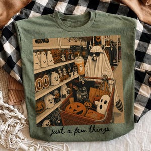 May include: An olive green t-shirt featuring a graphic of a raccoon wearing a ghost costume and sunglasses while pushing a shopping cart filled with Halloween decor, pumpkins, and a drink. The text "just a few things" is printed below the graphic.