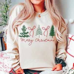 May include: Cream-coloured sweatshirt with a Christmas design. It features green Christmas trees and red bows, with the words "Merry Christmas" in red script. A festive and comfortable holiday jumper.