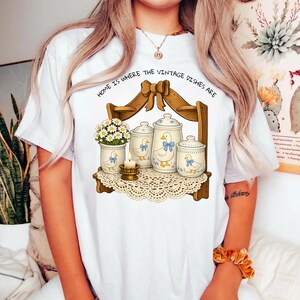 Op de afbeelding: Wit T-shirt met een vintage illustratie. Het ontwerp toont een houten plank met potten, een vaas met bloemen en een kaars. De potten hebben een eendenontwerp met blauwe strikken. De tekst "HOME IS WHERE THE VINTAGE DISHES ARE" staat erboven.