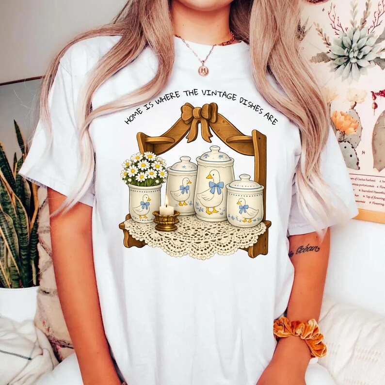 Op de afbeelding: Wit T-shirt met een vintage illustratie. Het ontwerp toont een houten plank met potten, een vaas met bloemen en een kaars. De potten hebben een eendenontwerp met blauwe strikken. De tekst "HOME IS WHERE THE VINTAGE DISHES ARE" staat erboven.