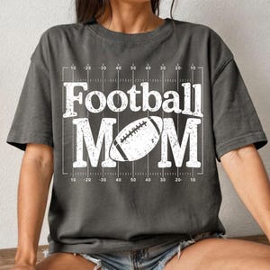 May include: Dark gray t-shirt with the words "Football MOM" in large white letters. A white football graphic is centered within the word "MOM". The shirt has a football field design with yard lines.