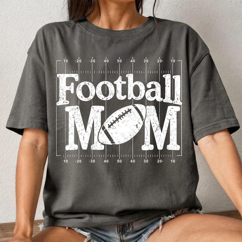 May include: Dark gray t-shirt with the words "Football MOM" in large white letters. A white football graphic is centered within the word "MOM". The shirt has a football field design with yard lines.