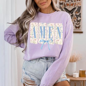 May include: A light purple sweatshirt with the word "AMEN" in blue block letters. The letters are set against a floral background, and a blue bow is below the word. The sweatshirt is paired with denim shorts.