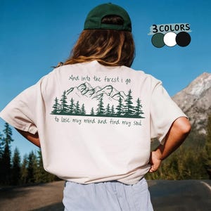 以下が含まれることがあります： 緑の山と森のグラフィックと「And into the forest I go to lose my mind and find my soul」というテキストが入ったベージュのTシャツ。画像には、緑、白、黒の3つのカラーオプションも表示されています。
