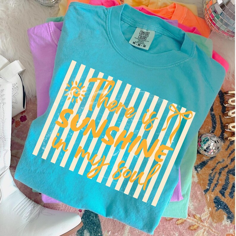 Puede incluir: Una pila de camisetas Comfort Colors coloridas. La camiseta superior es turquesa con el texto "There is sunshine in my soul" en amarillo. El texto est&aacute; sobre un fondo a rayas blancas. Otras camisetas son rosas, moradas y naranjas.