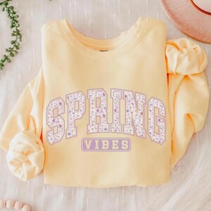May include: A light yellow sweatshirt with the word "SPRING" in large, lavender block letters filled with a floral pattern. Below, the word "VIBES" is in a lavender rectangle. The sweatshirt is folded on a white surface.