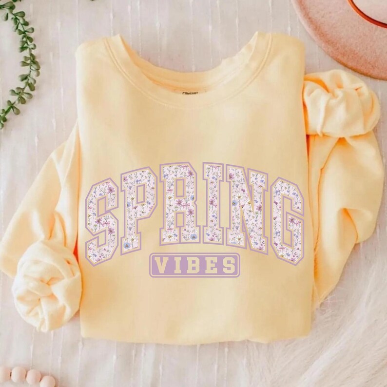 May include: A light yellow sweatshirt with the word "SPRING" in large, lavender block letters filled with a floral pattern. Below, the word "VIBES" is in a lavender rectangle. The sweatshirt is folded on a white surface.